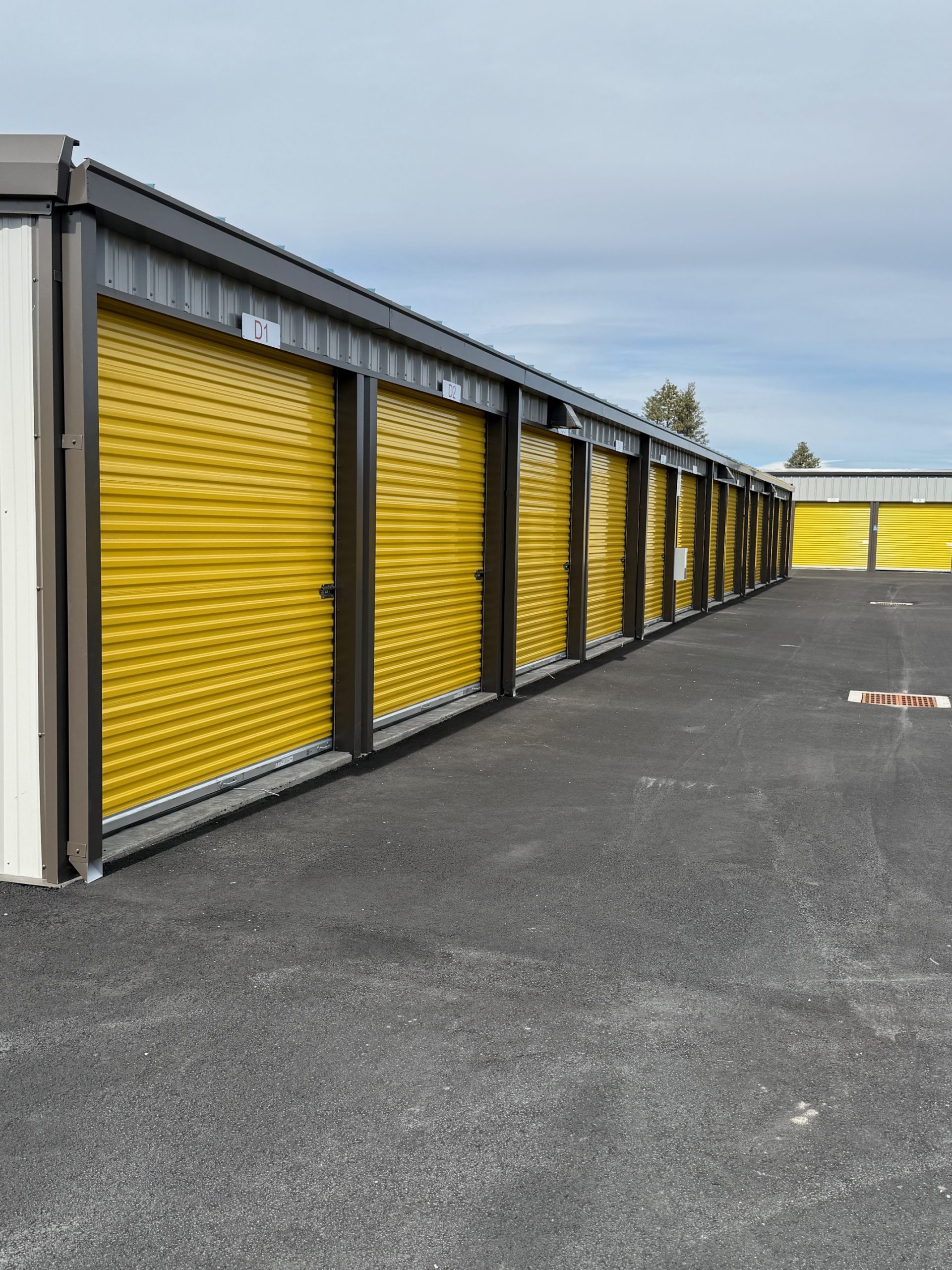 A row of storage units with closed yellow roll-up doors on a paved surface under a cloudy sky.