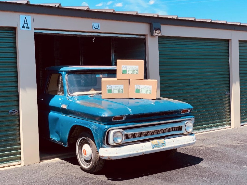 A vintage blue pickup truck is parked halfway out of a storage unit with three cardboard boxes labeled "STORAGE" stacked on its hood.