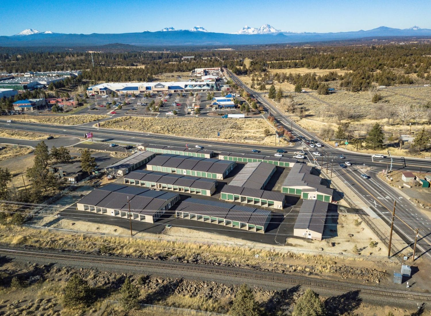 Aerial view of a large self-storage facility with multiple buildings near a road intersection, surrounded by dry terrain, shops, and distant mountains.