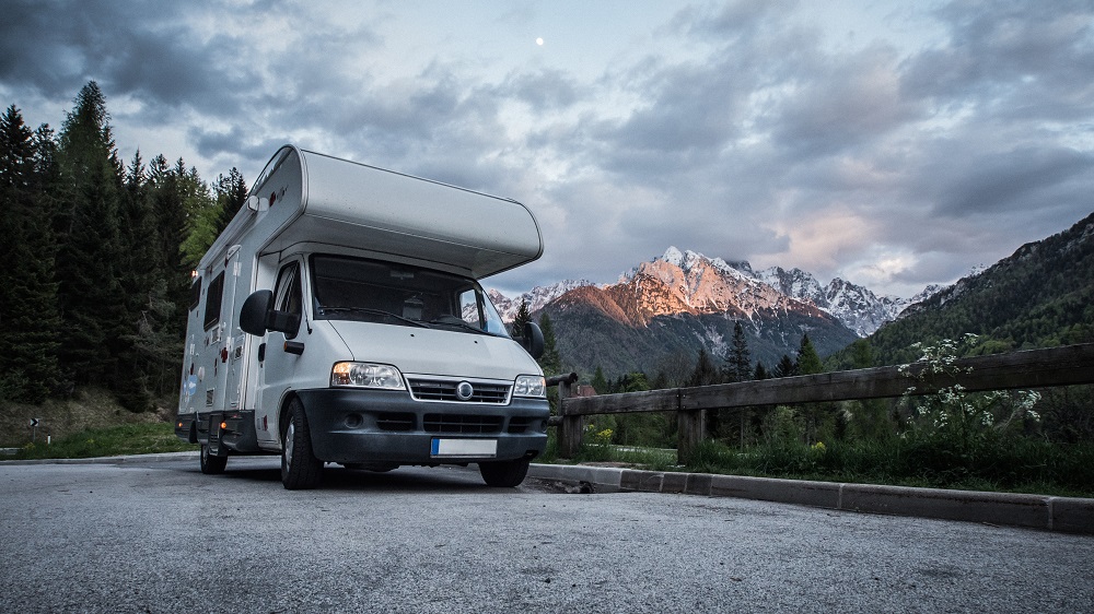 A white camper van is parked on a roadside near a wooden fence with forest and snow-capped mountains in the background under a cloudy sky.