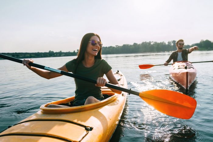 Two people kayaking on a calm lake, each in a yellow kayak, smiling and paddling under a clear sky.