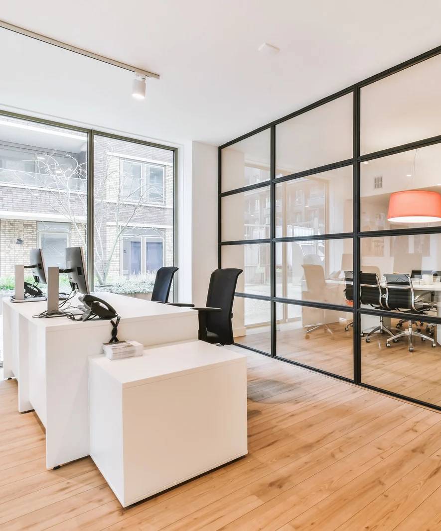 Modern office space with two white desks, black chairs, computers, wooden floor, large windows, and a glass-walled meeting room in the background.