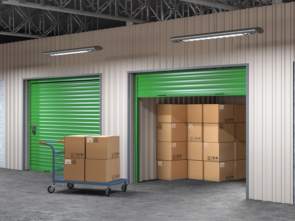 A storage unit with its green door open, revealing stacked cardboard boxes inside. A cart with more boxes is positioned nearby on the concrete floor.
